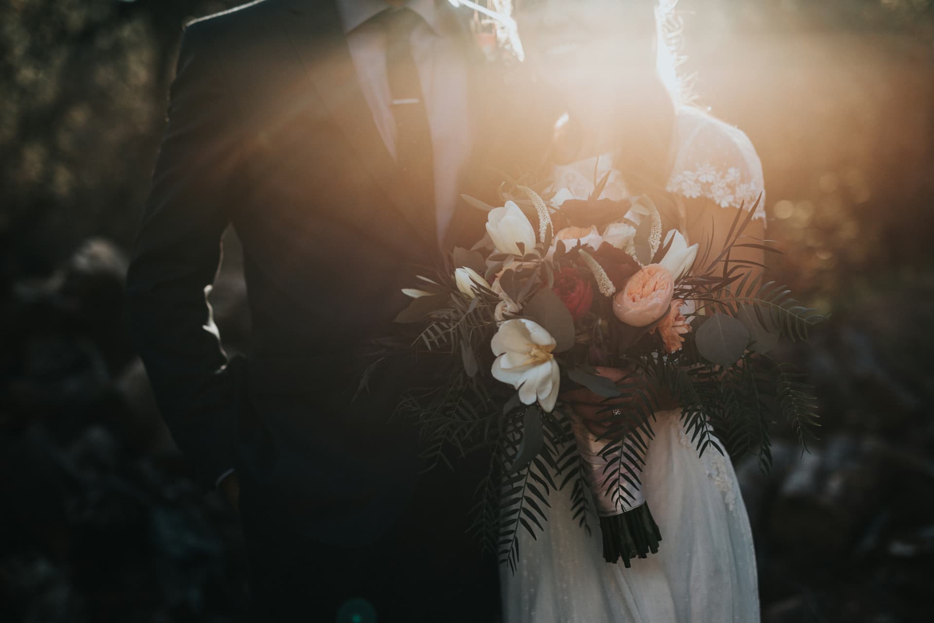 Bride and groom holding a bouquet of roses and ferns in golden backlight during their wedding ceremony
