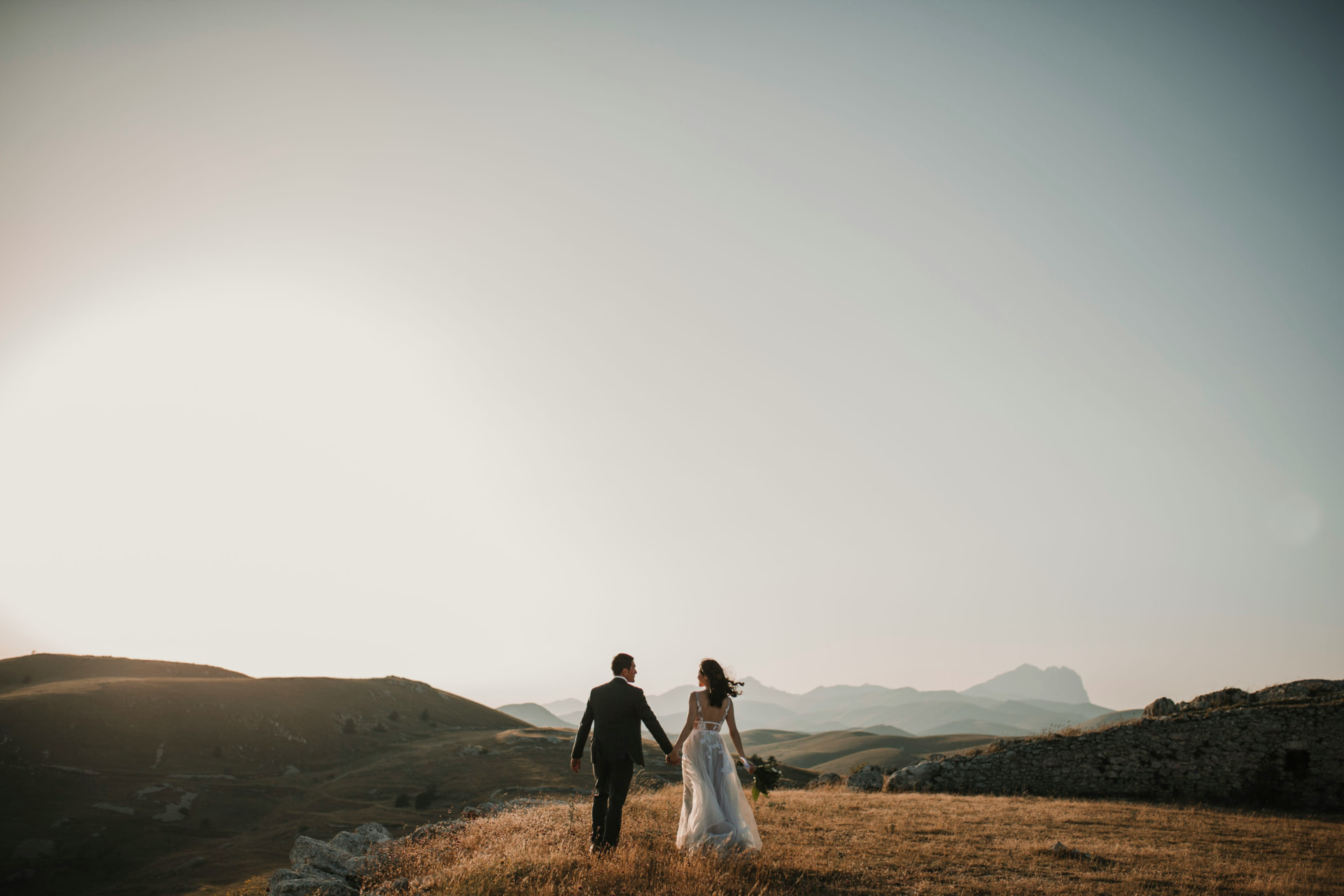 Newlywed couple walking hand in hand across golden hills at sunset